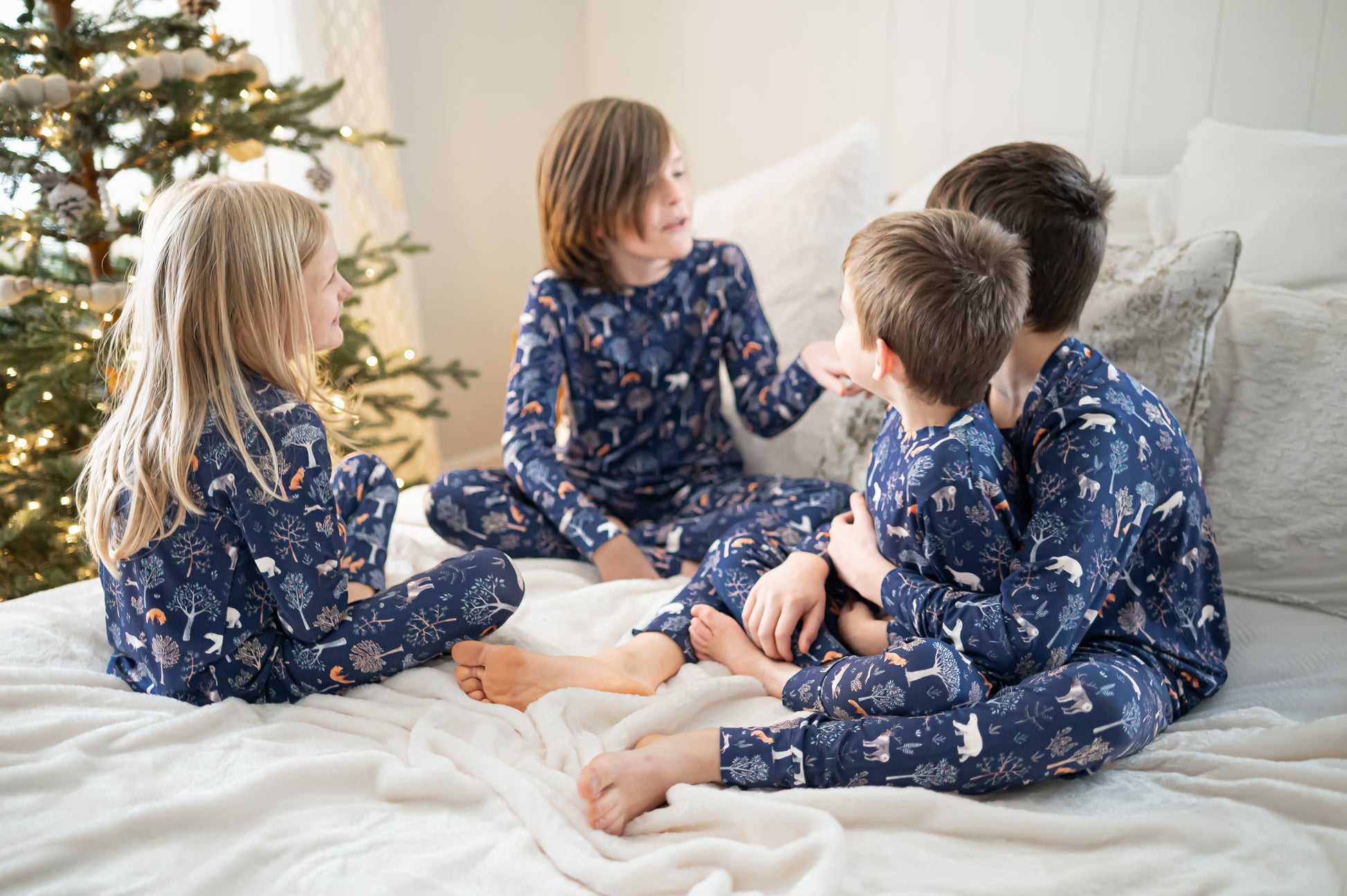 Four children in matching pajamas sitting on a bed with a Christmas tree in the background