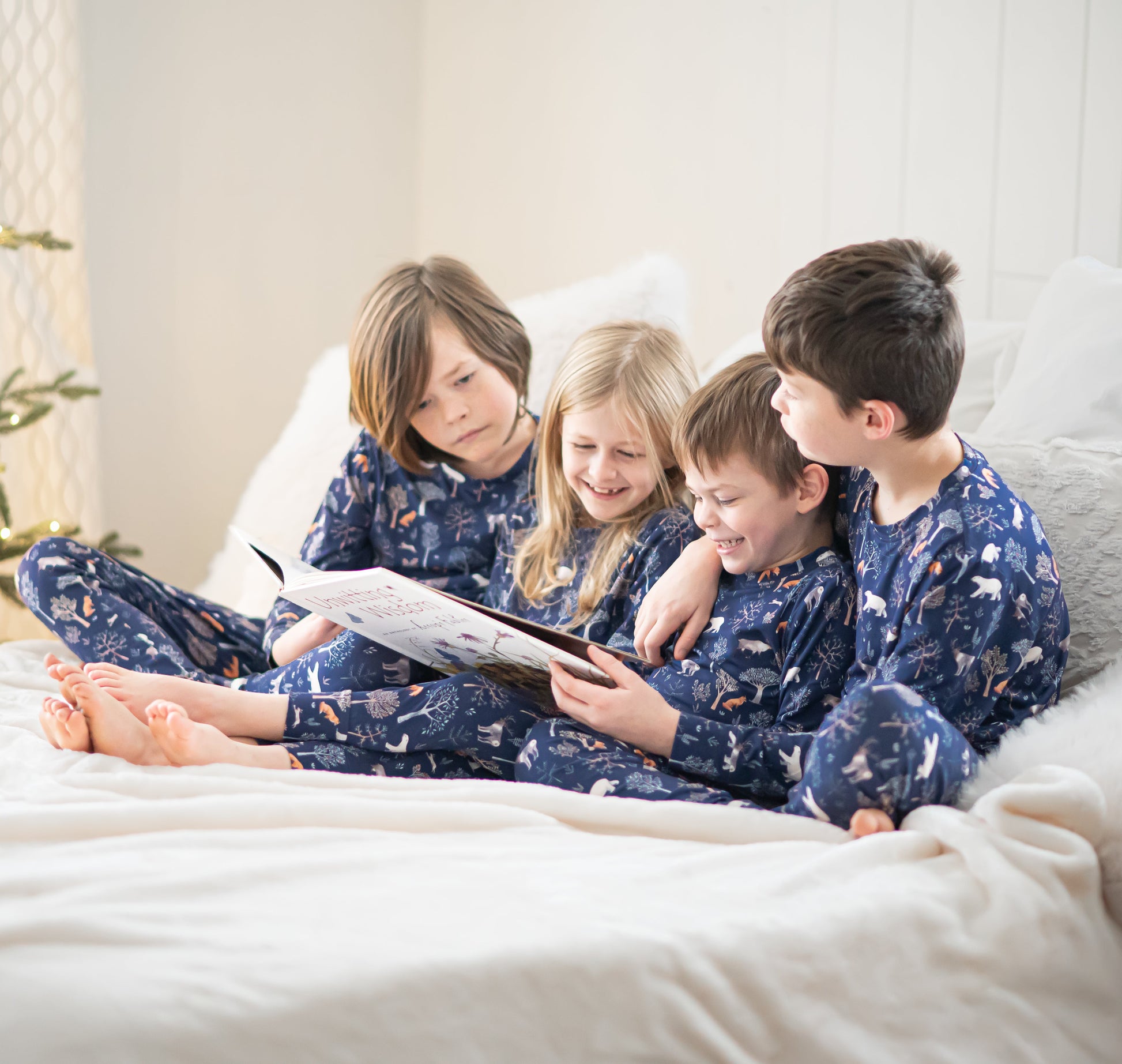Four children in matching pajamas sitting on a bed reading a book, with a decorated Christmas tree in the background.