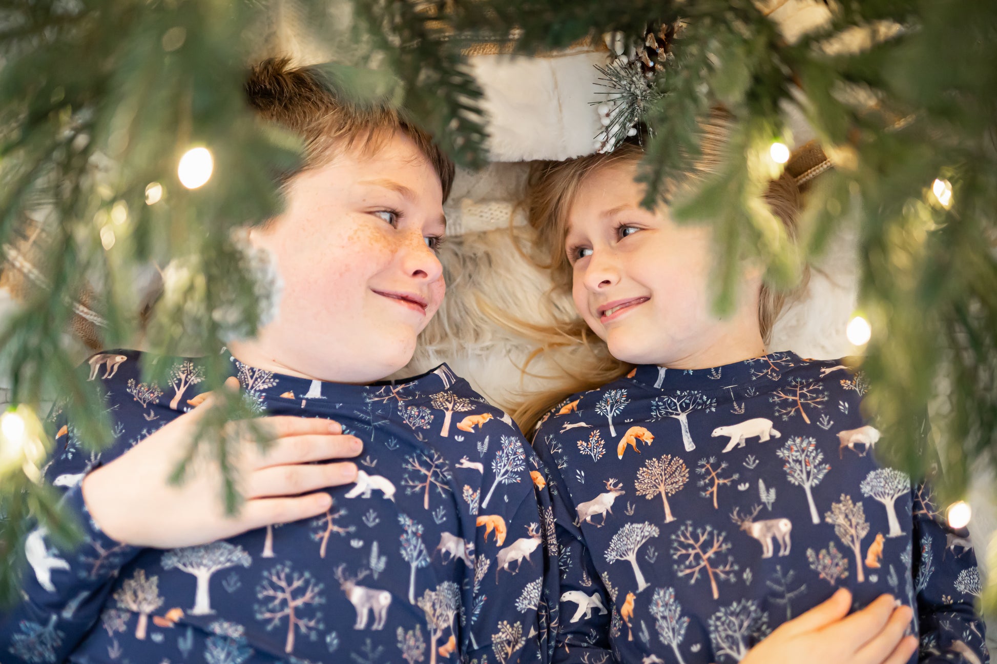 Two children wearing matching pajamas with a forest pattern, lying under a Christmas tree.