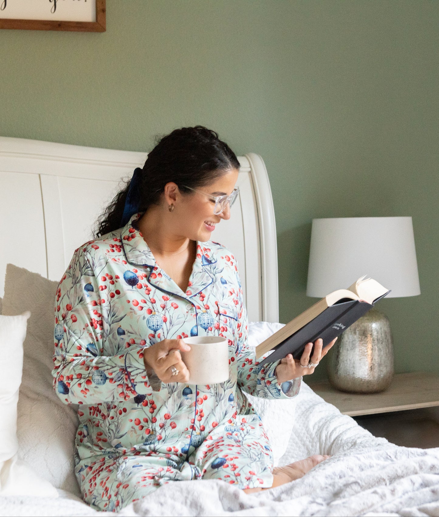 Woman in floral pajamas reading a book on a bed in a cozy bedroom.