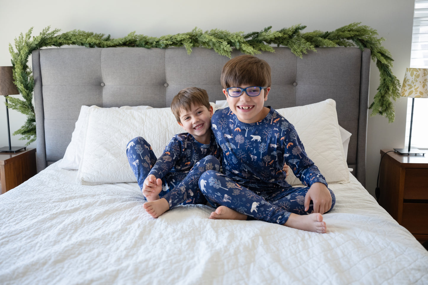 Two children in matching pajamas sitting on a bed with a festive headboard.