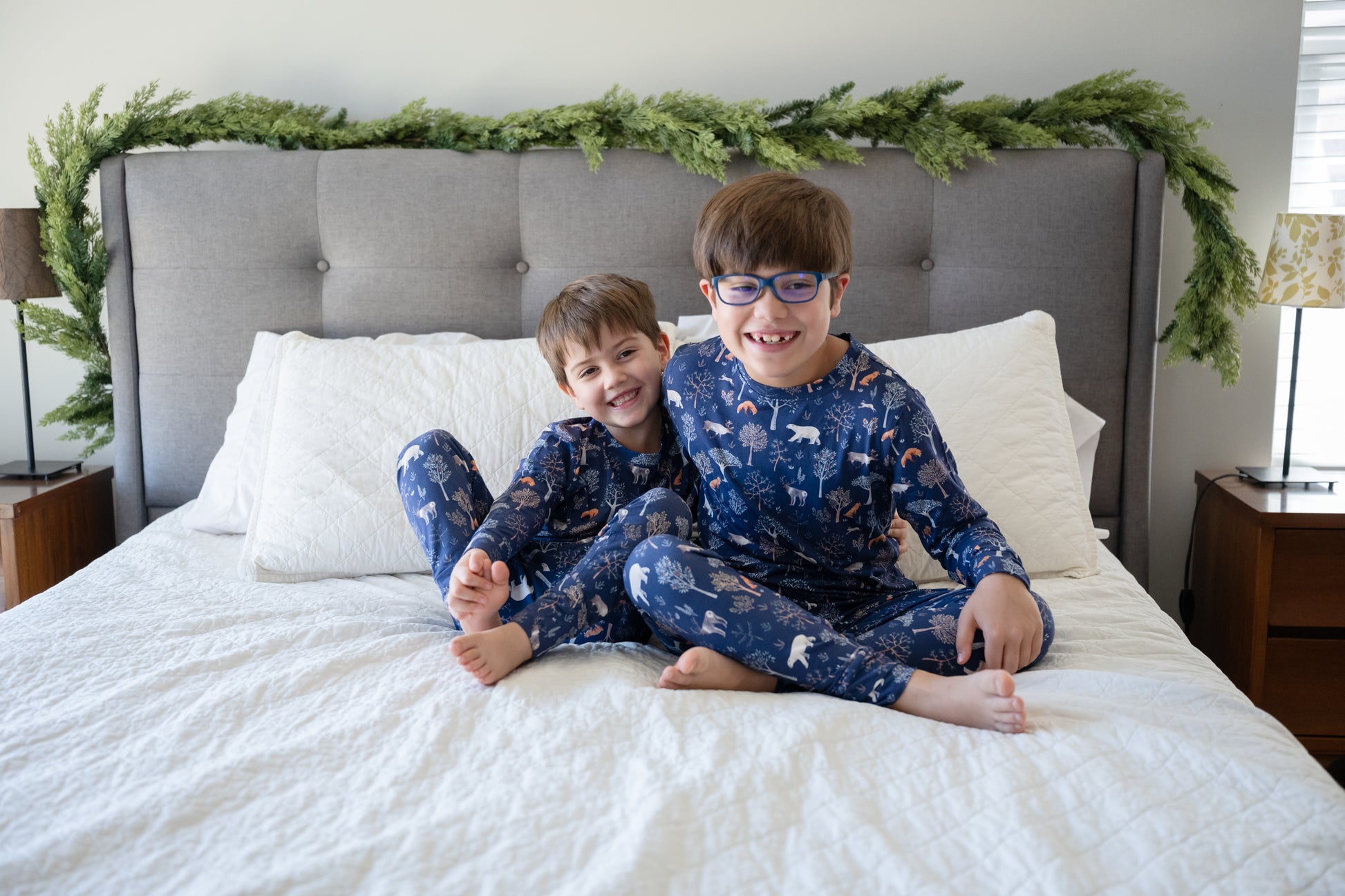 Two children in matching pajamas sitting on a bed with a festive headboard.